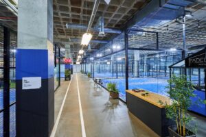 Binnenzicht van een moderne padelhal aan de Johan Huizingalaan in Amsterdam met meerdere blauwe padelbanen en een glazen wand.